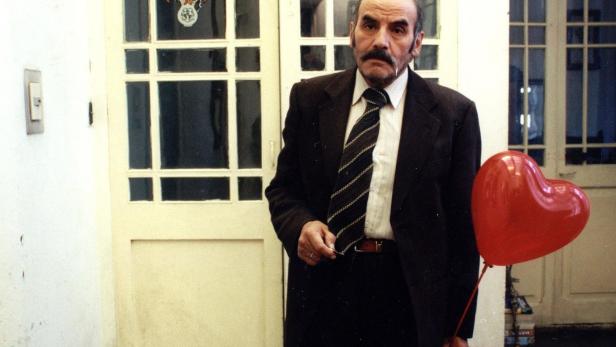 Ein Mann mit Anzug und rotem Herzluftballon steht vor einer Tür.