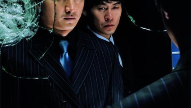 Zwei Männer in Anzügen stehen hinter einem zerbrochenen Spiegel.