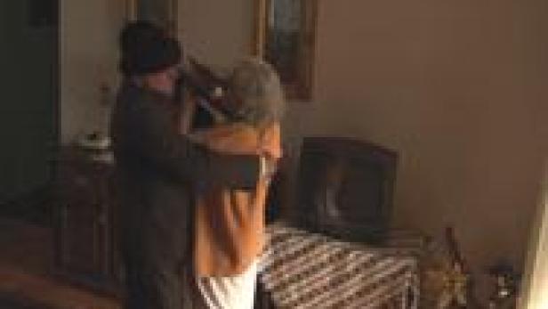 Ein Mann mit Mütze umarmt eine ältere Frau in einem Wohnzimmer.