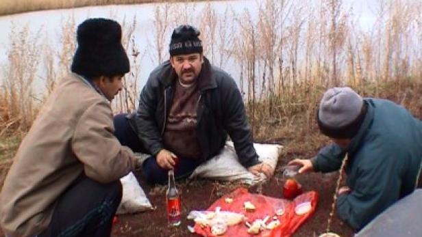 Drei Männer sitzen am Ufer eines Sees und trinken Wodka und essen Snacks.