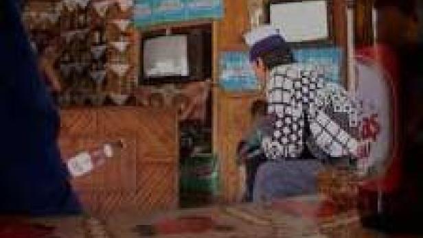 Ein Mann mit Mütze sitzt in einem kleinen Raum mit Fernseher.