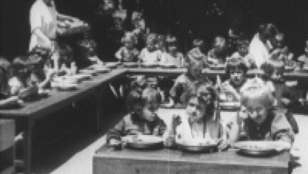 Eine Gruppe von Kindern sitzt an Tischen und isst in einer Art Freiluft-Cafeteria.