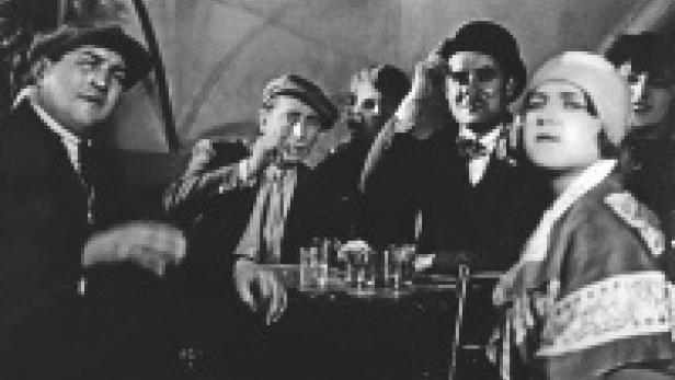 Eine Gruppe von Menschen sitzt an einem Tisch mit Schnapsgläsern in einem verrauchten Raum.