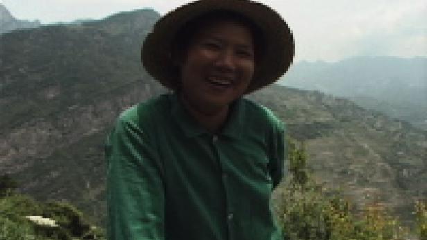 Eine lächelnde Frau mit Hut steht vor einer Berglandschaft.