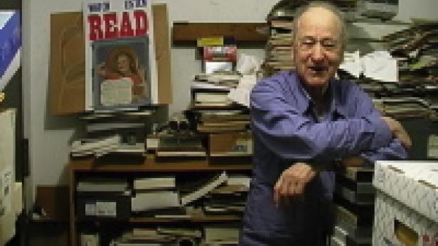 Ein älterer Mann steht vor einem Stapel Kartons in einem mit Büchern und Papieren gefüllten Raum.