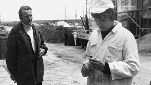 Zwei Männer stehen auf einer Baustelle, einer im Anzug, der andere in Arbeitskleidung.