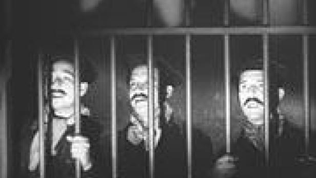 Drei Männer mit Hüten und falschen Bärten stehen hinter Gittern.