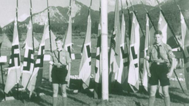 Zwei Jungen in Uniform stehen vor einer Reihe von Flaggen mit Hakenkreuzen.