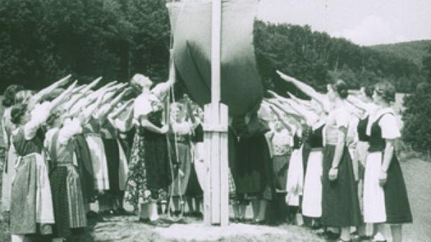 Eine Gruppe junger Frauen in Tracht salutiert vor einem Fahnenmast.