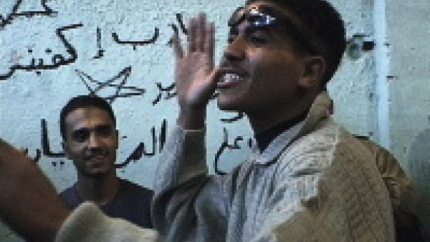 Ein junger Mann gestikuliert vor einer Wand mit arabischer Schrift.