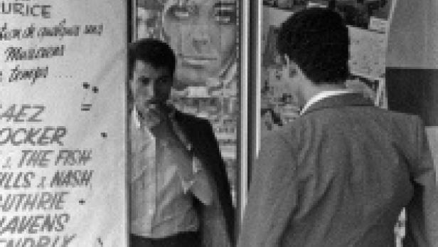 Ein Mann mit Schnurrbart steht vor einem Plakat, während eine andere Person vorbeigeht.