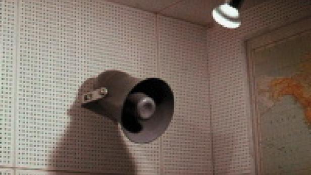 Ein grauer Lautsprecher hängt an einer Wand mit einer Weltkarte im Hintergrund.