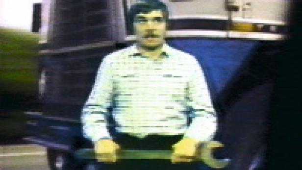 Ein Mann mit Schnurrbart hält einen großen Schraubenschlüssel vor einem Bus.