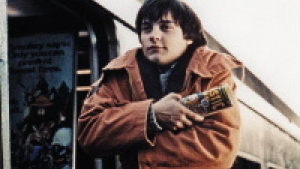 Ein junger Mann in einer orangefarbenen Jacke steht mit verschränkten Armen vor einem Zug.