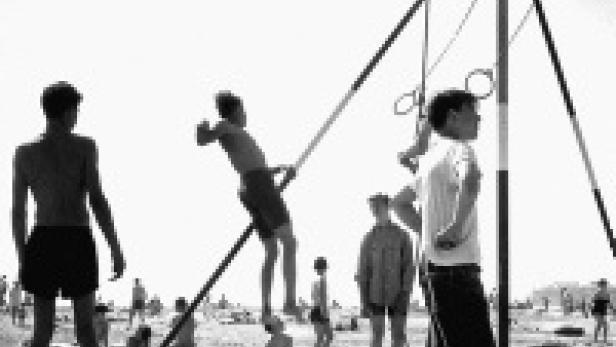 Eine Gruppe junger Männer turnt an einem improvisierten Reck auf einem sonnigen Strand.