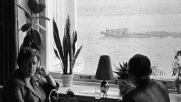 Zwei Personen sitzen an einem Tisch vor einem Fenster mit Blick auf ein Gewässer und ein Schiff.