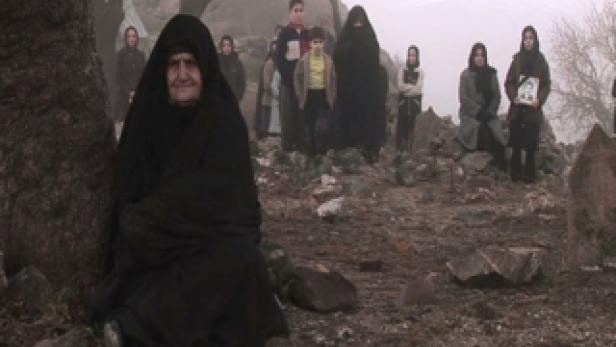 Eine ältere Frau in traditioneller Kleidung sitzt vor einer Gruppe von Menschen auf einem Friedhof.