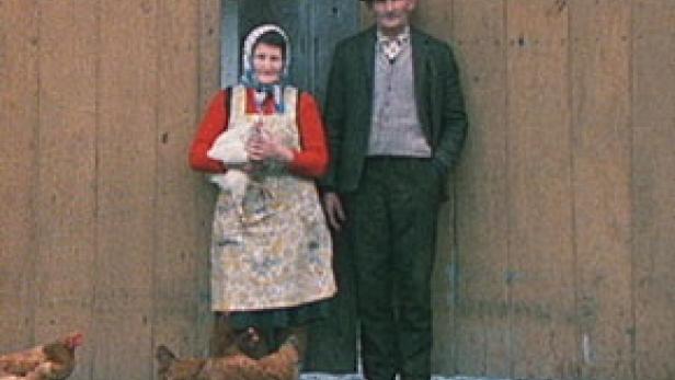 Ein älteres Paar steht vor einer Holzwand, die Frau hält ein Huhn, während Hühner um sie herum laufen.