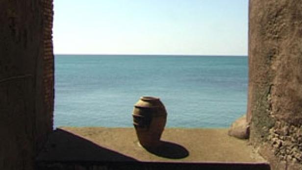 Ein Tongefäß steht vor dem blauen Meer, gesehen durch eine steinerne Fensteröffnung.