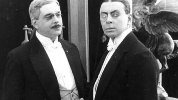 Zwei Männer in Anzügen, einer mit Brille und Schnurrbart, der andere mit starrem Blick.