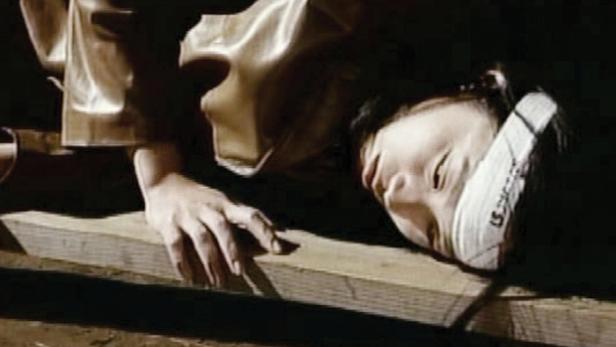 Eine verletzte Frau mit einem Verband liegt auf einem Holzbalken.