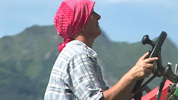 Eine Frau mit Kopftuch fährt einen Traktor vor einer Bergkulisse.