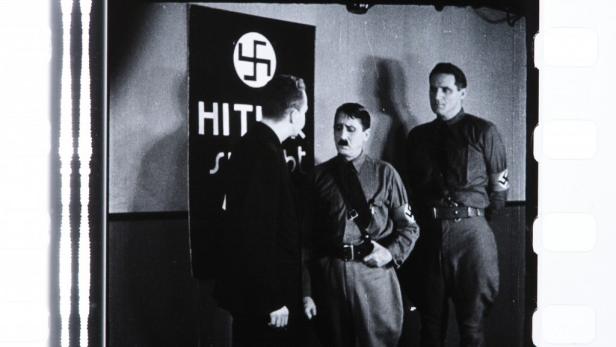 Eine Schwarzweißaufnahme von Adolf Hitler mit zwei Männern vor einem Hakenkreuz-Schild.