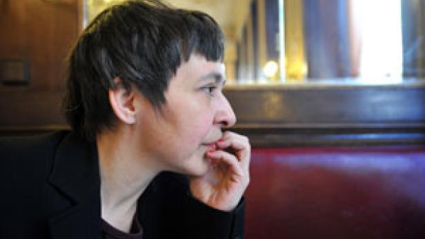 Eine Frau mit dunkler Jacke sitzt in einem Café und blickt nach rechts.