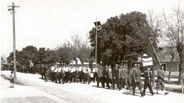Ein Schwarzweißfoto einer Parade mit Hakenkreuzflagge und Bannern in einer Stadt.