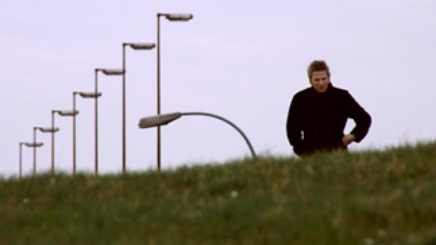 Ein Mann steht auf einem grasbewachsenen Hügel, im Hintergrund eine Reihe von Straßenlaternen.