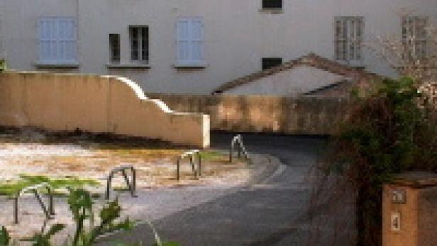 Ein asphaltierter Weg führt an einer Mauer und einem Gebäude vorbei.