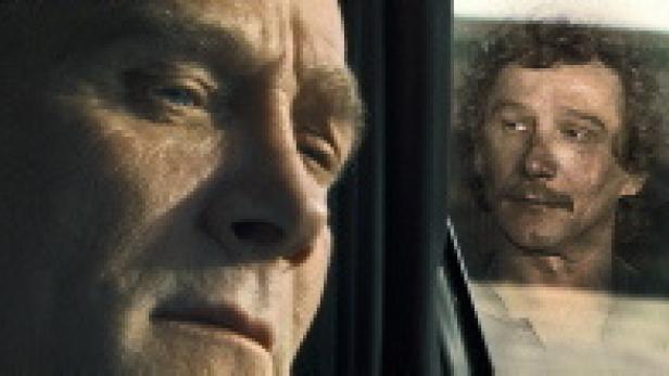 Ein Mann blickt ernst, während ein anderer im Autofenster zu sehen ist.