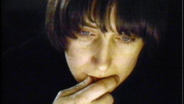 Eine Frau mit dunklen Haaren und hellen Augen blickt nach unten und berührt ihre Lippen.