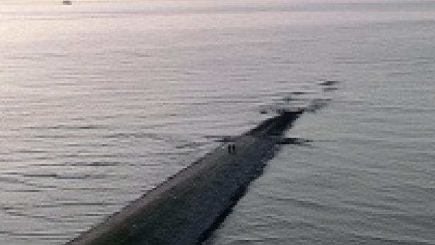 Ein langer Steg ragt ins Meer, am Horizont ist ein Schiff zu sehen.