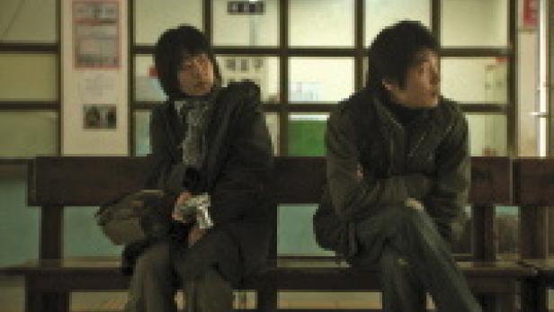 Zwei Männer sitzen auf einer Bank in einem Wartebereich und schauen in die Ferne.