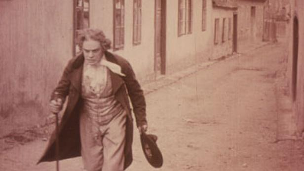 Ludwig van Beethoven geht mit einem Stock und einem Hut in der Hand eine Gasse entlang.