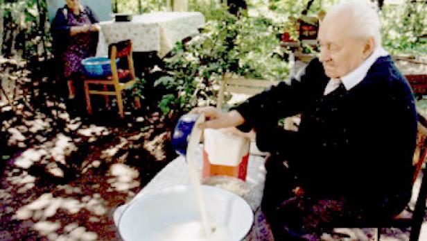 Ein älterer Mann gießt Milch in eine Schüssel im Freien, während eine Frau im Hintergrund steht.