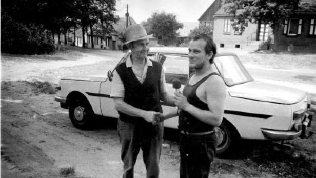 Ein Mann mit Hut schüttelt einem Reporter vor einem Auto die Hand.