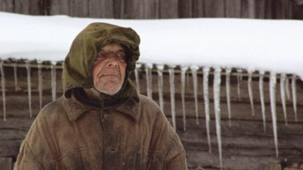 Ein älterer Mann mit wettergegerbtem Gesicht und Kapuze steht vor einem schneebedeckten Holzgebäude mit Eiszapfen.