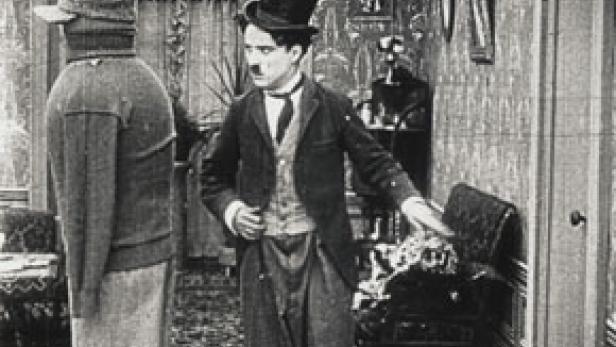 Charlie Chaplin in Anzug und Melone gestikuliert in einem Raum, in dem eine weitere Person steht.