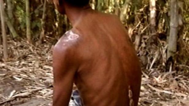 Ein Mann sitzt mit freiem Oberkörper in einem Waldstück.