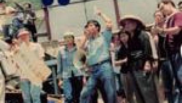 Eine Menschenmenge steht im Freien, einige gestikulieren und halten Schilder hoch.