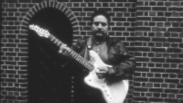 Ein Mann mit Gitarre posiert vor einer Backsteinmauer.