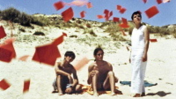 Drei Personen sitzen und stehen an einem Strand, während rote Papierstücke in der Luft schweben.