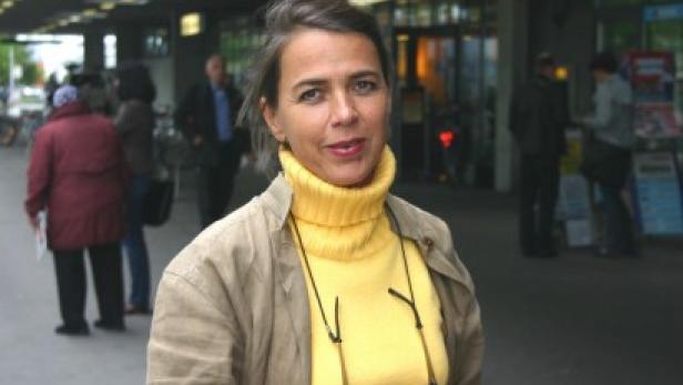 Eine Frau mit gelbem Rollkragenpullover und Brille steht auf einem Bürgersteig.