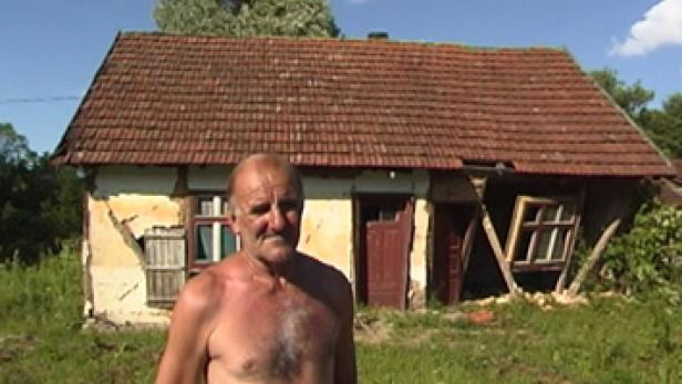 Ein älterer Mann steht vor einem beschädigten Haus mit Rissen in den Wänden.