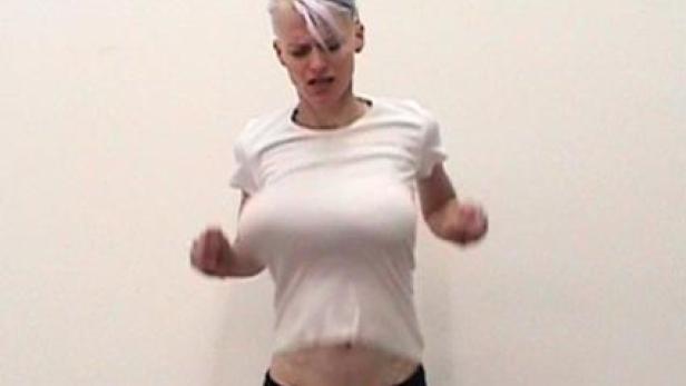 Eine Frau mit kurzen, blonden Haaren und einem weißen T-Shirt tanzt vor einer weißen Wand.