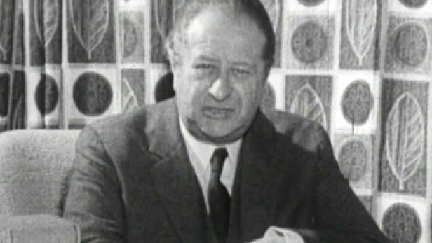 Ein Mann im Anzug sitzt vor einem gemusterten Vorhang.