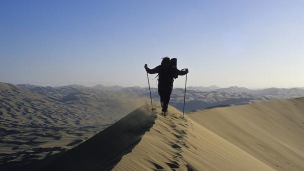 Ein Wanderer mit Rucksack und Stöcken geht über eine Sanddüne.