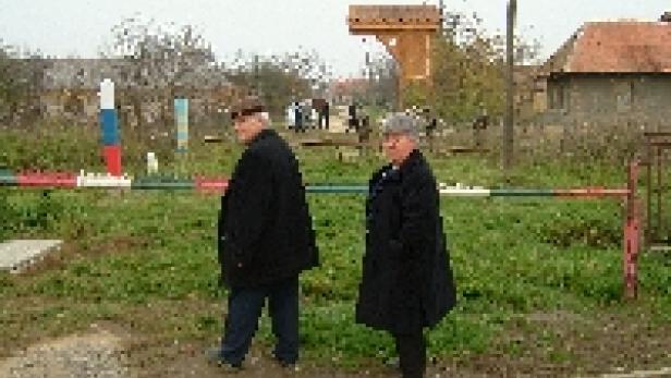Zwei ältere Menschen stehen an einem Bahnübergang in einer ländlichen Gegend.